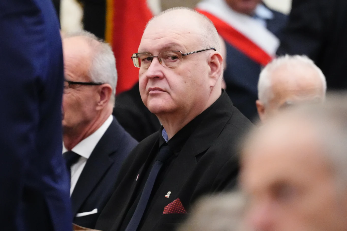 Starszy mężczyzna w garniturze i okularach siedzi wśród innych osób podczas oficjalnego wydarzenia, skupiony patrzy w stronę aparatu.