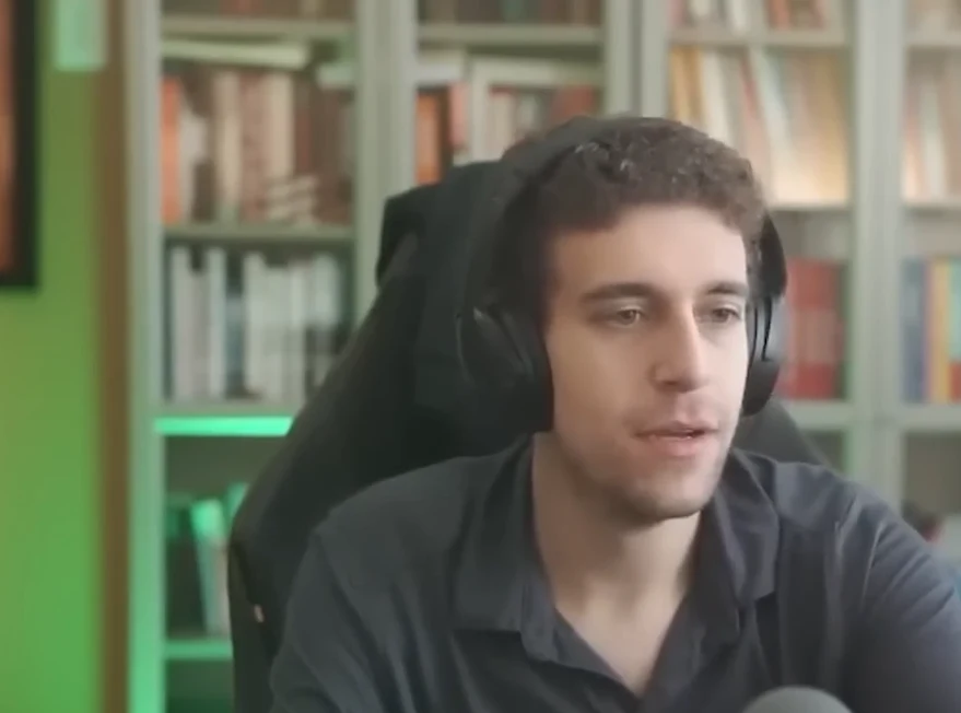 Daniel Naroditsky Mężczyzna z krótkimi, kręconymi włosami, w ciemnej koszuli, siedzi na krześle przed regałem z książkami i ma na głowie duże słuchawki.