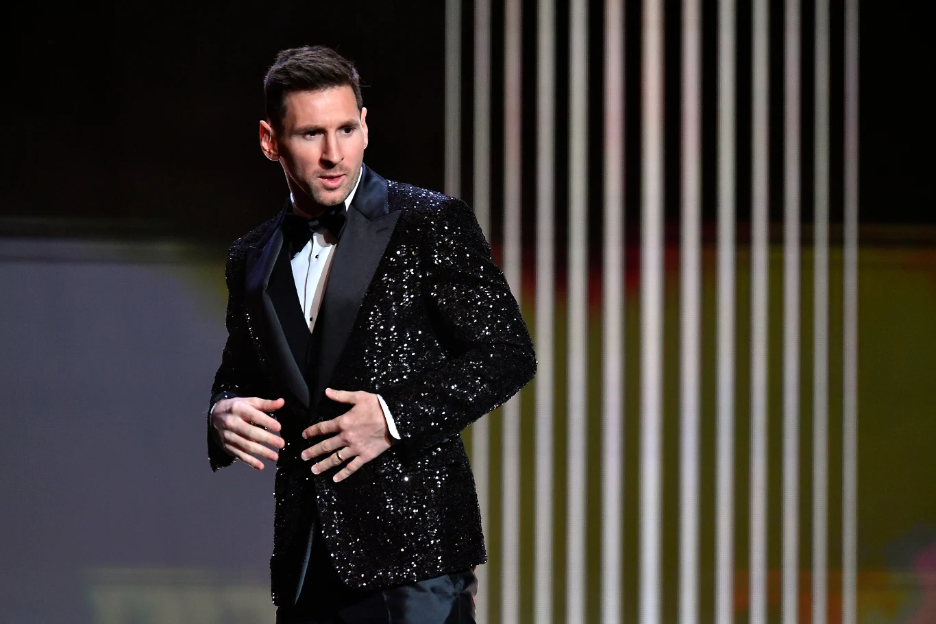 Lionel Messi, 29.11.2021 r. Mężczyzna w czarnym cekinowym garniturze i muchą stoi na scenie oświetlonej pionowymi białymi światłami w tle.