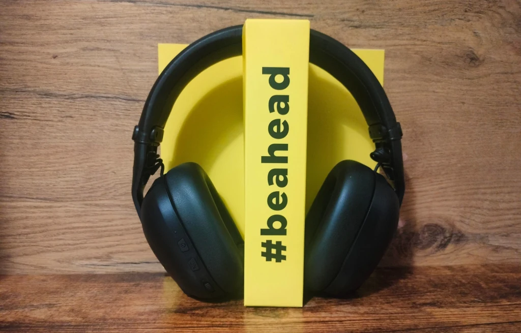 Czarne, duże słuchawki nauszne ustawione na tle żółtego pudełka z widocznym napisem #beahead, całość umieszczona na drewnianym podłożu, dominuje kontrast żółci i czerni.