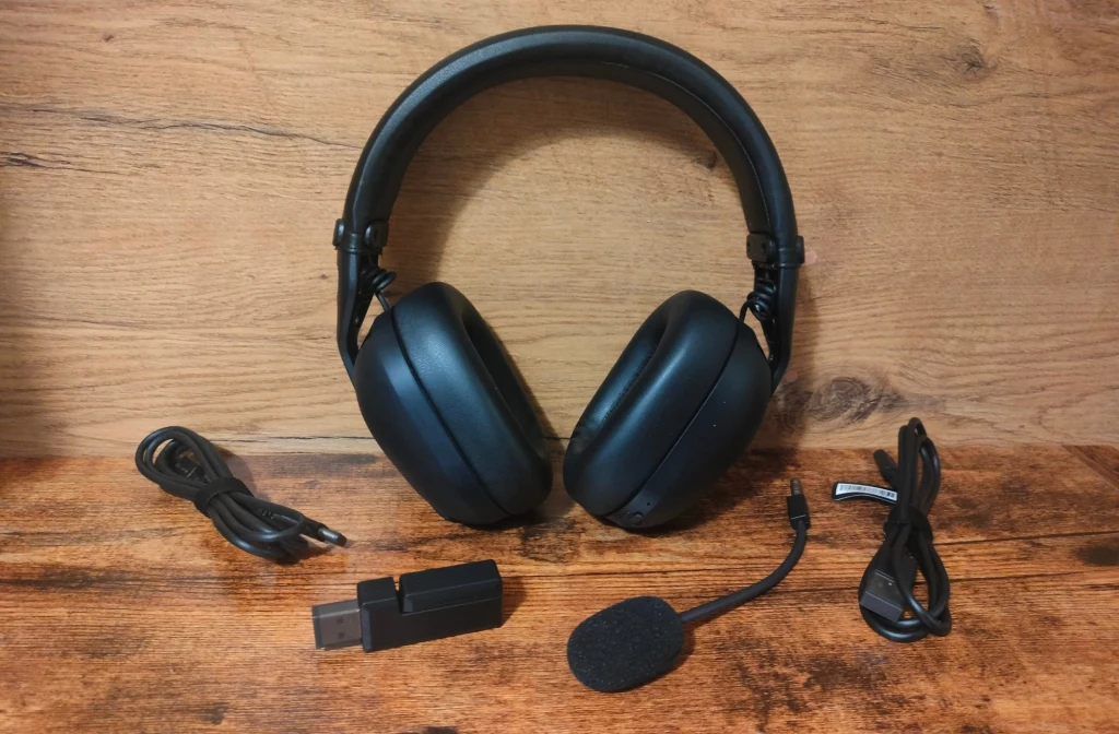 Czarne słuchawki nausznie wraz z odłączanym mikrofonem, donglem USB i dwoma różnymi kablami leżą na drewnianej powierzchni.