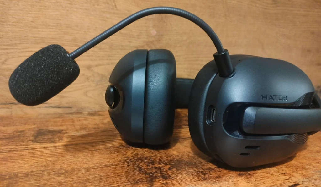 Czarne słuchawki nauszne z mikrofonem na wysięgniku leżą na drewnianej powierzchni, widoczny port ładowania oraz marka Hator na jednej z muszli.