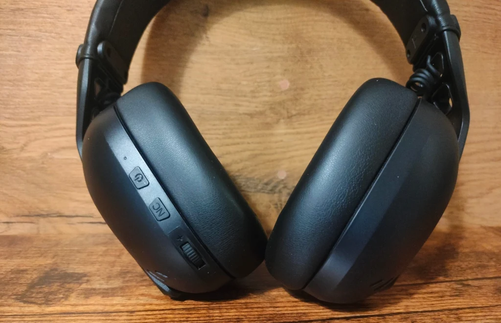 Czarne bezprzewodowe słuchawki nauszne z widocznymi przyciskami sterującymi na lewej muszli, o matowym wykończeniu, położone na drewnianej powierzchni.