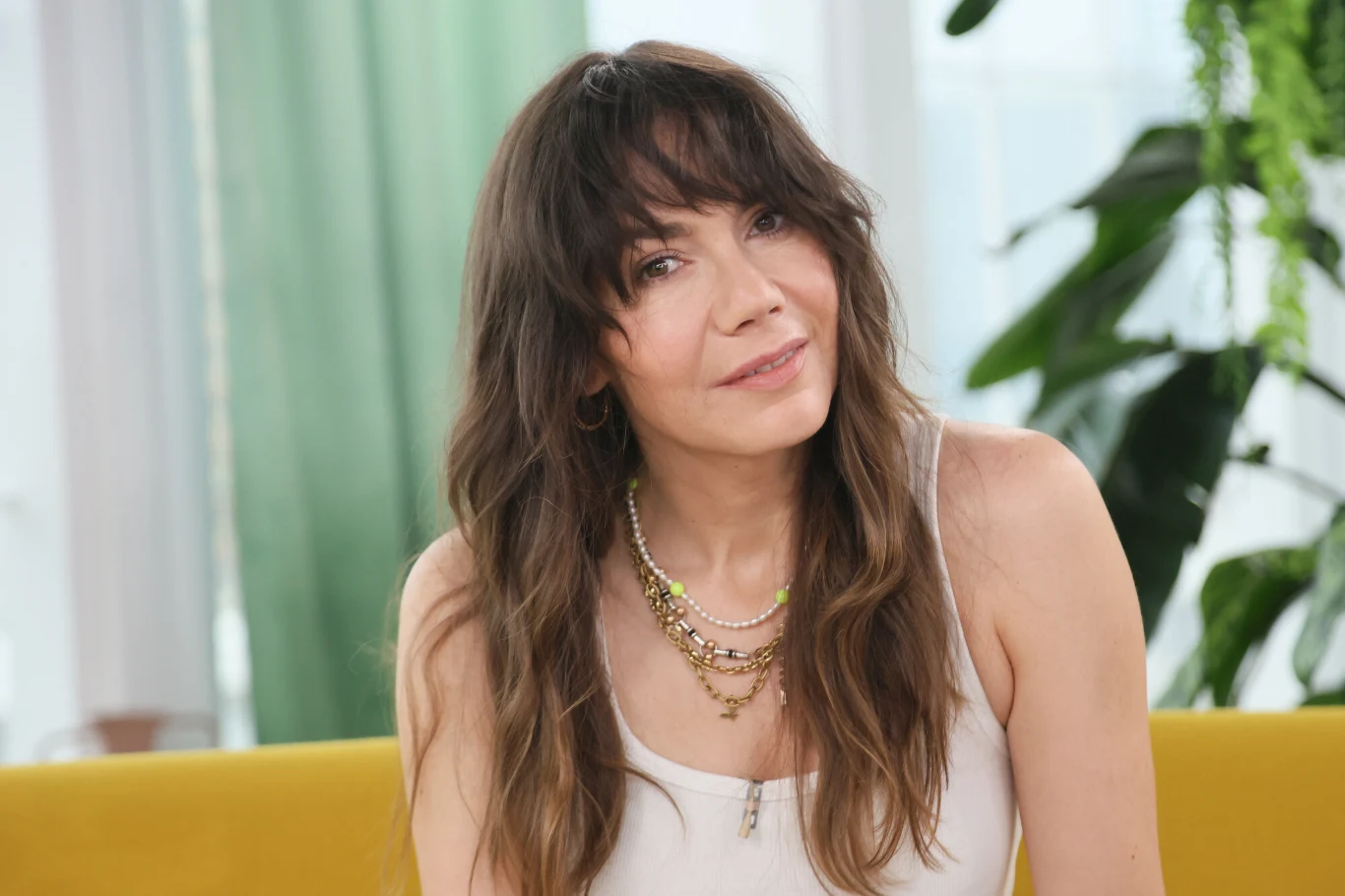 Olga Bołądź Kobieta z długimi, brązowymi włosami i grzywką w białym topie, siedząca na żółtej sofie. Na szyi nosi kilka złotych i srebrnych naszyjników, w tle zielone rośliny doniczkowe oraz okno z białymi zasłonami.