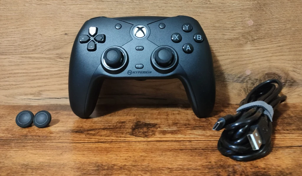 Hyperkin Competitor Czarny kontroler do gier z dwiema gałkami analogowymi, przyciskami funkcyjnymi i krzyżakiem, po lewej stronie dwie dodatkowe nakładki na gałki, po prawej kabel USB z przewiązaną opaską, całość na tle drewnianej powierzchni.