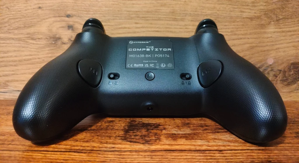 Hyperkin Competitor Czarny kontroler do gier z widocznymi przyciskami i informacjami producenta leżący na drewnianej powierzchni.