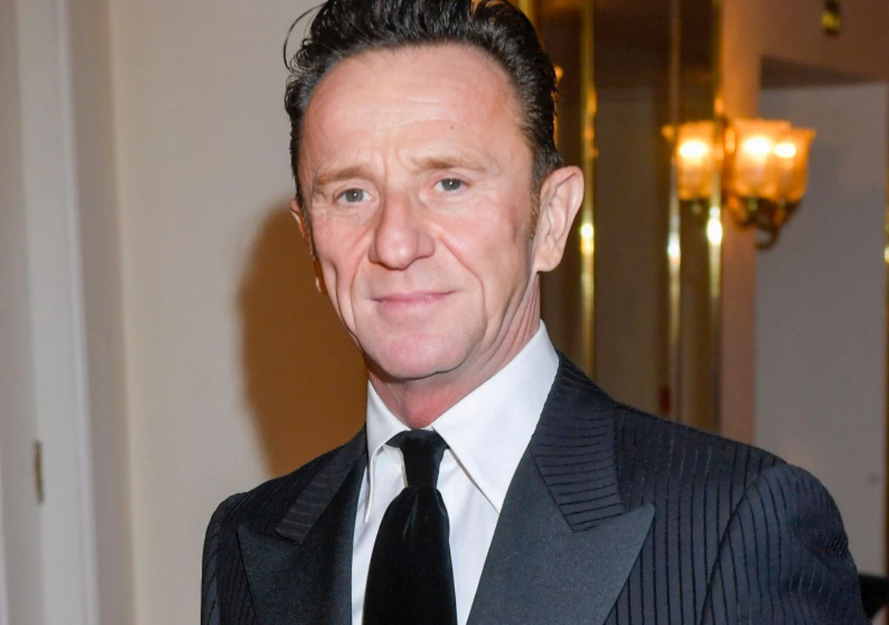 Paweł Wilczak Paweł Wilczak w garniturze z białą koszulą i czarnym krawatem, stojący w eleganckim, dobrze oświetlonym wnętrzu ze złocistymi lampami ściennymi w tle.