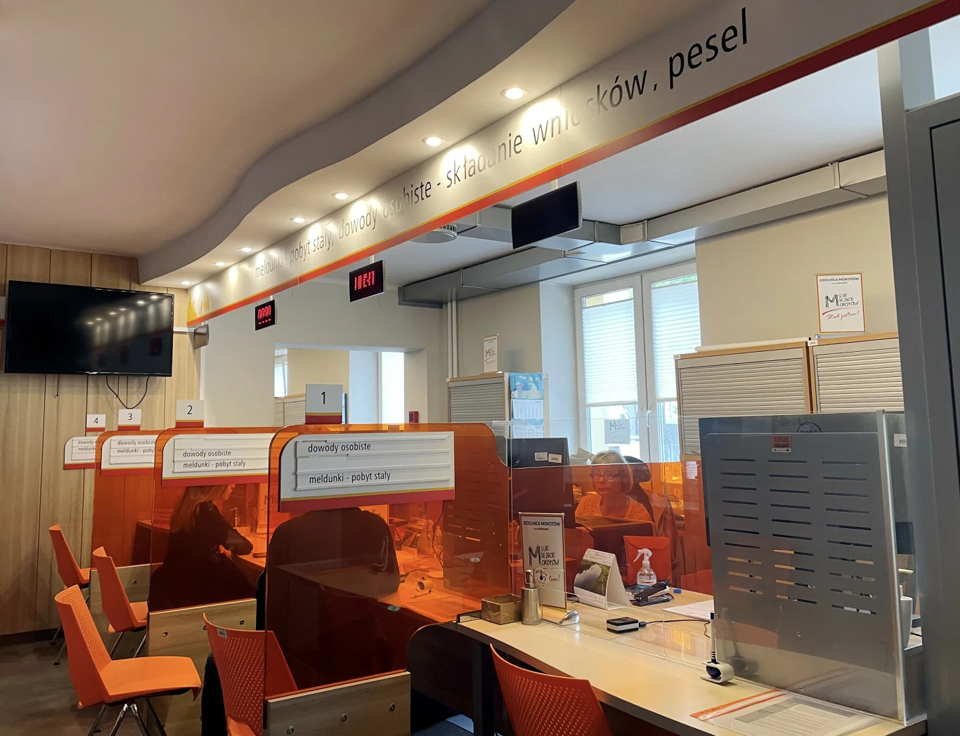 Dowód osobisty jest ważny przez 10 lat. Stanowiska obsługi klienta z pomarańczowymi przegrodami i numeracją, krzesła dla interesantów, ekrany z informacją o kolejce oraz tabliczki wskazujące zakres obsługiwanych spraw.