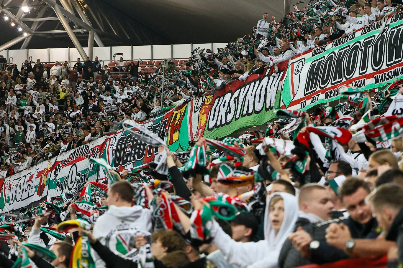 Kibice Legii Warszawa Tłum kibiców piłkarskich z szalikami i flagami w barwach klubu, trybuny pełne entuzjazmu, transparenty z nazwami dzielnic Warszawy, dominują kolory biały, zielony i czerwony.
