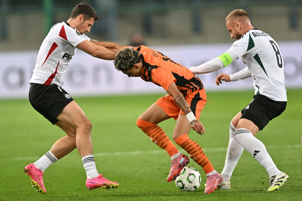 Szachtar Donieck - Legia Warszawa Dwóch piłkarzy w białych strojach walczy o piłkę z zawodnikiem w pomarańczowym trykocie, akcja toczy się w dynamicznym tempie na zielonym boisku, widoczne są koncentracja i fizyczny kontakt podczas meczu piłki nożnej.