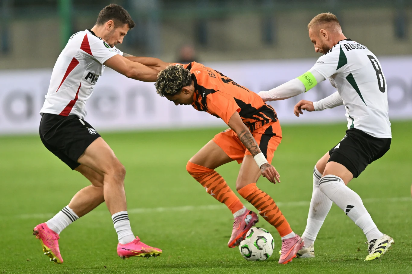 Szachtar Donieck - Legia Warszawa Dwóch piłkarzy w białych strojach walczy o piłkę z zawodnikiem w pomarańczowym trykocie, akcja toczy się w dynamicznym tempie na zielonym boisku, widoczne są koncentracja i fizyczny kontakt podczas meczu piłki nożnej.