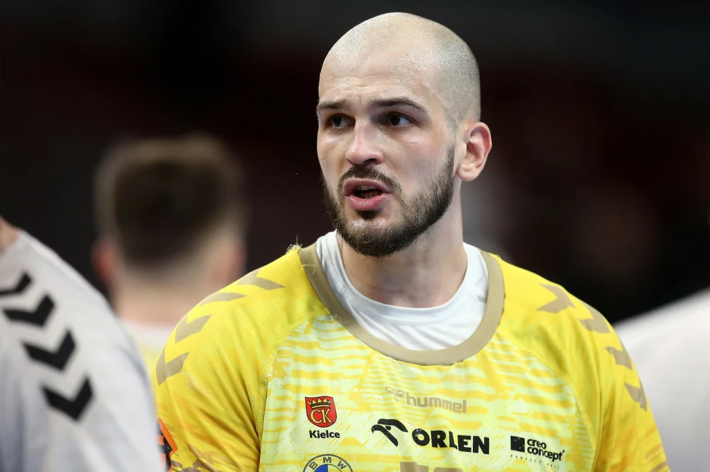 Zawodnik piłki ręcznej w żółtej koszulce z logotypami klubu oraz sponsorów, o wyrazistym wyrazie twarzy, w trakcie rozgrywki sportowej z rozmytym tłem i innymi postaciami w tle.