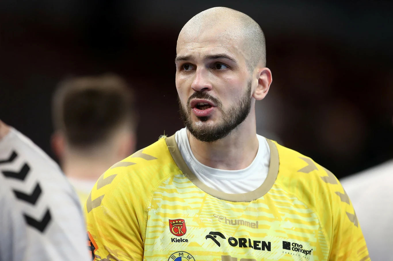 Zawodnik piłki ręcznej w żółtej koszulce z logotypami klubu oraz sponsorów, o wyrazistym wyrazie twarzy, w trakcie rozgrywki sportowej z rozmytym tłem i innymi postaciami w tle.