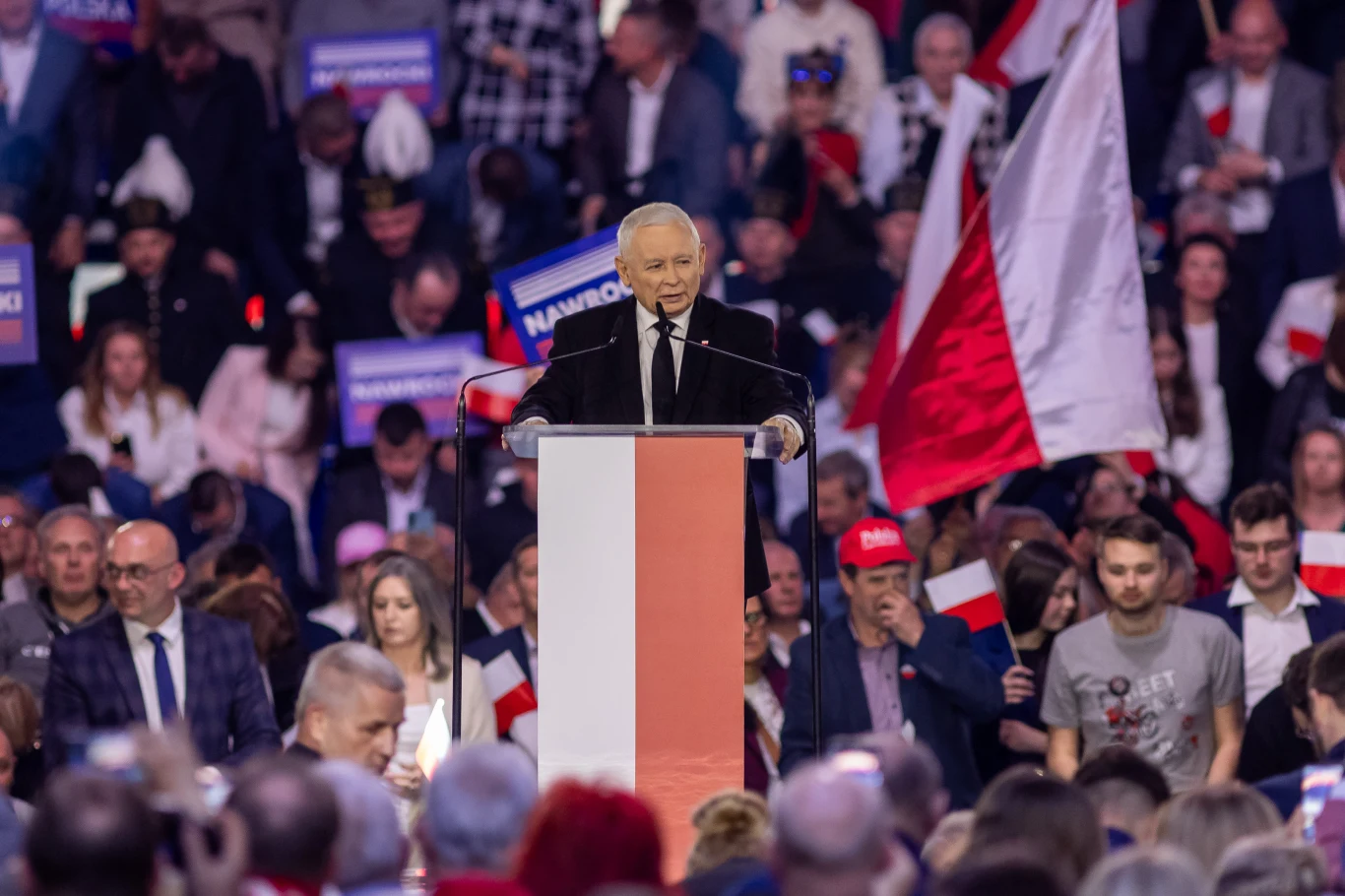 Konwencja programowa PiS składa się z 17 sesji głównych i ponad stu paneli specjalistycznych Polityk przemawia z mównicy udekorowanej biało-czerwoną flagą, w tle tłum ludzi z flagami Polski i transparentami, atmosfera skupiona, dominują barwy narodowe.