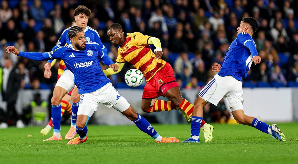 Afimico Pululu osaczony przez defensorów RC Strasbourg Dwóch piłkarzy w niebieskich strojach próbuje odebrać piłkę zawodnikowi w żółto-czerwonym stroju, podczas meczu piłkarskiego na zielonej murawie. W tle widać kibiców na trybunach.