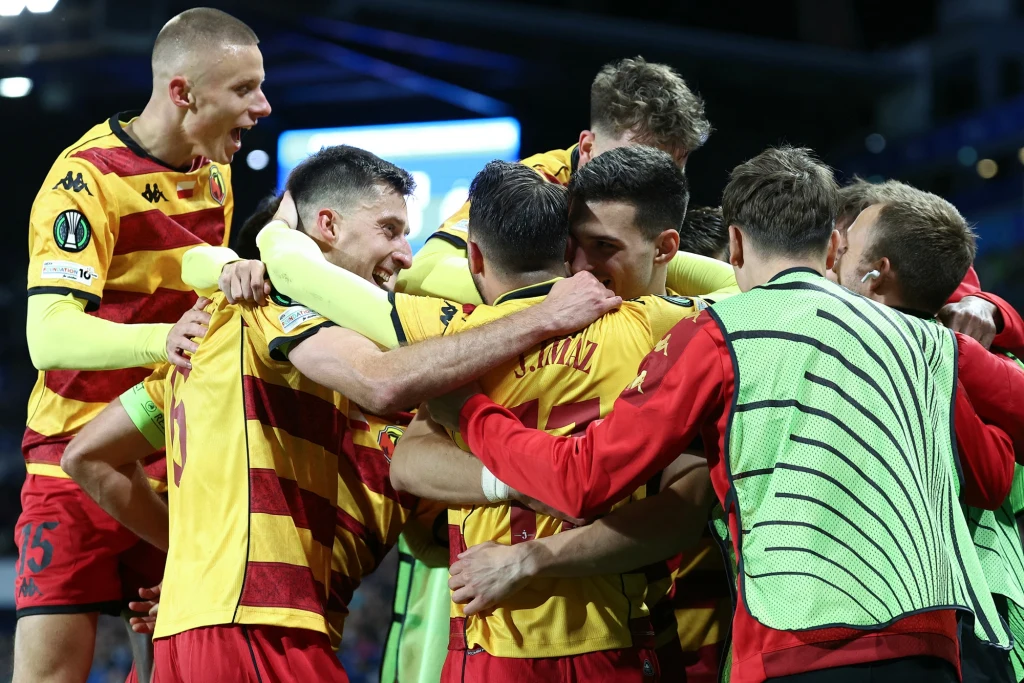 W tym momencie Jagiellonia prowadziła w gościnie 1:0 Grupa piłkarzy w żółto-czerwonych strojach ściska się i cieszy z sukcesu na boisku podczas meczu, na ich twarzach widać radość i sportowe emocje.