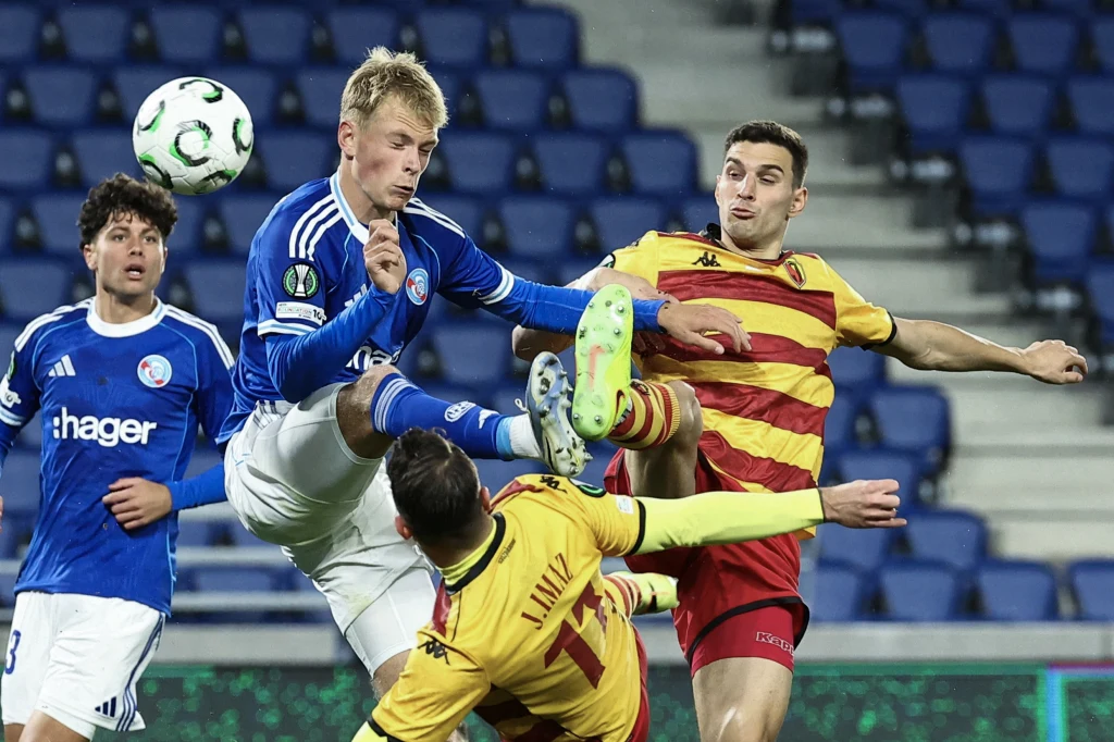 W starciu lidera Ekstraklasy z trzecią drużyną Ligue 1 nikt nie odstawiał nogi Intensywna rywalizacja piłkarzy w trakcie meczu piłki nożnej, zawodnik w niebieskiej koszulce próbuje przejąć piłkę wysokim kopnięciem, otoczony przez graczy w żółto-czerwonych i niebieskich strojach, wszyscy skoncentrowani na akcji. Stadion wypełniają...