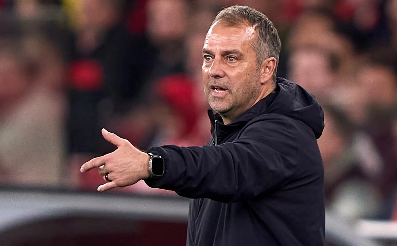 Hansi Flick Mężczyzna w średnim wieku, ubrany w ciemną bluzę, energicznie gestykuluje stojąc na tle rozmytej widowni stadionu sportowego.