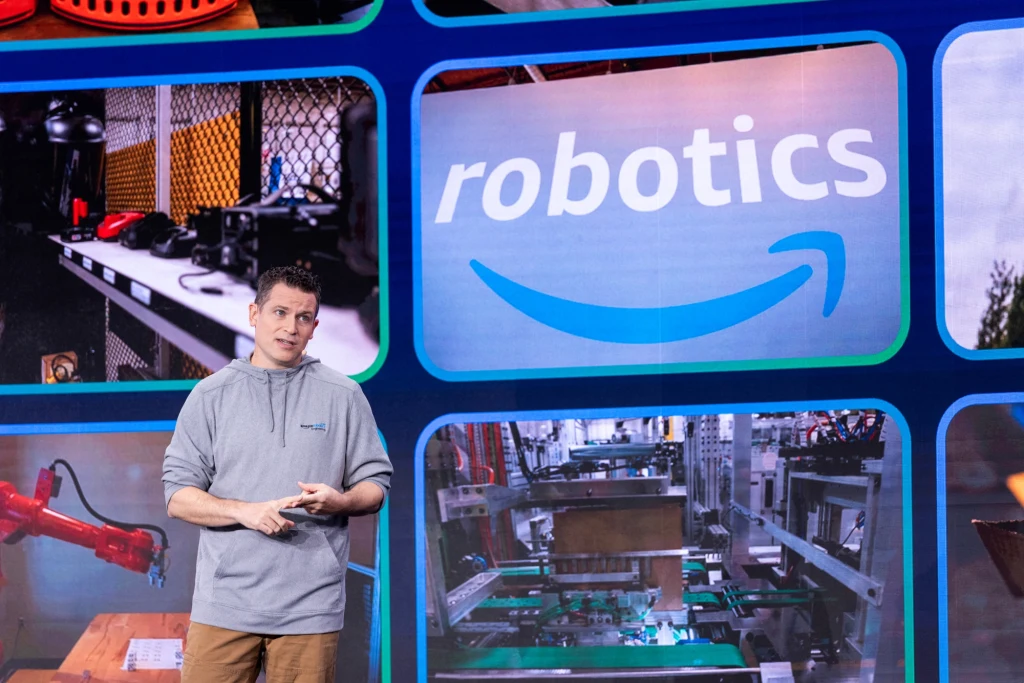 Mężczyzna w szarej bluzie prezentuje tematy związane z robotyką, a tło stanowią zdjęcia pokazujące maszyny, roboty i duży napis 'robotics' z logotypem Amazon.