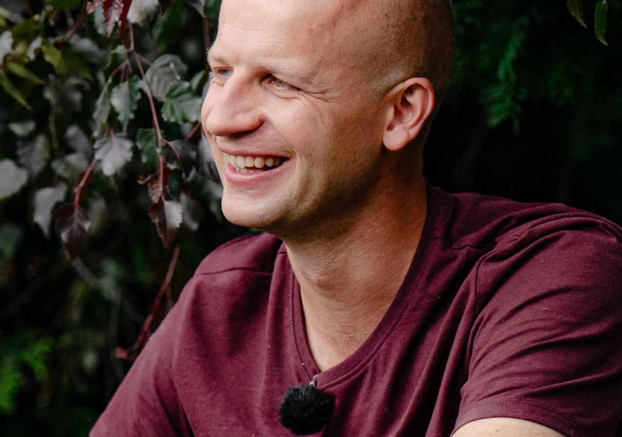 Gabriel z "Rolnik szuka żony" Uśmiechnięty Gabriel z programu "Rolnik szuka żony" w bordowej koszulce siedzi na tle zielonych liści, z mikrofonem przypiętym do koszulki.