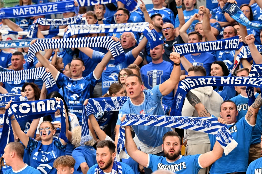 Tłum kibiców ubranych w niebieskie koszulki i szaliki z nadrukiem Wisła Płock, unoszących ręce i kibicujących z pasją, niektórzy śpiewają lub skandują.
