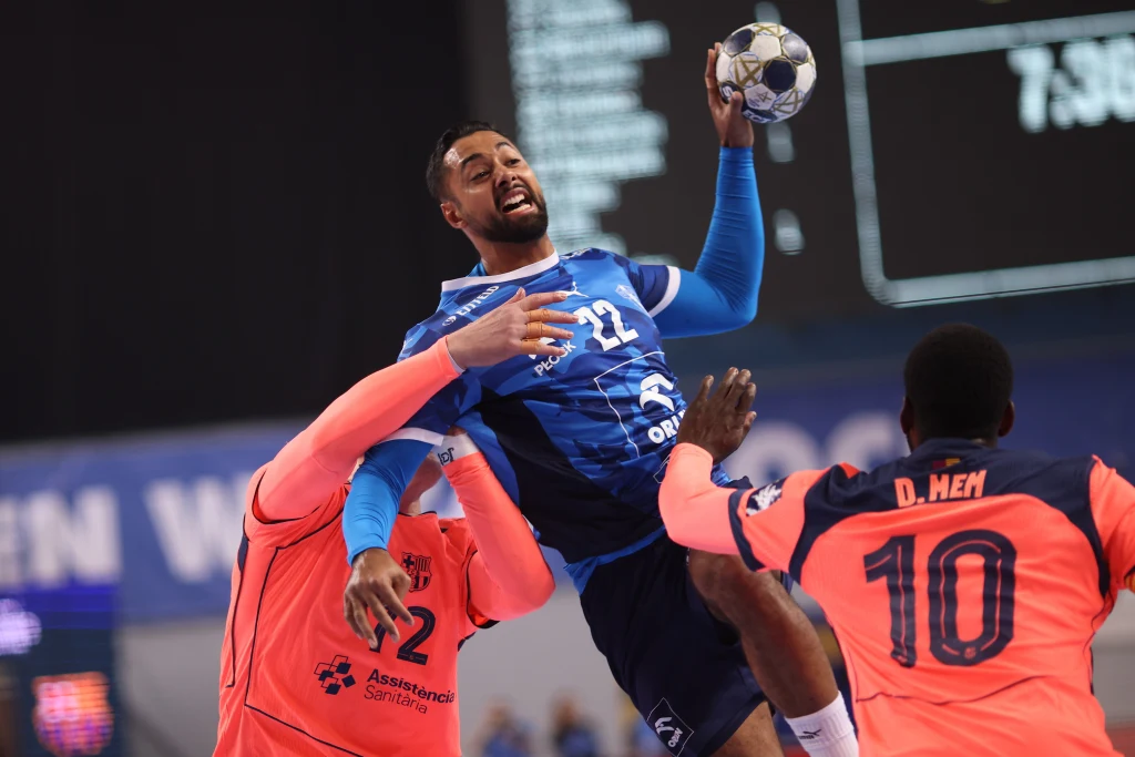 Zawodnik w niebieskim stroju skacze z piłką do ręki podczas meczu piłki ręcznej, starając się oddać rzut pomimo obrony ze strony dwóch przeciwników ubranych w pomarańczowe koszulki.