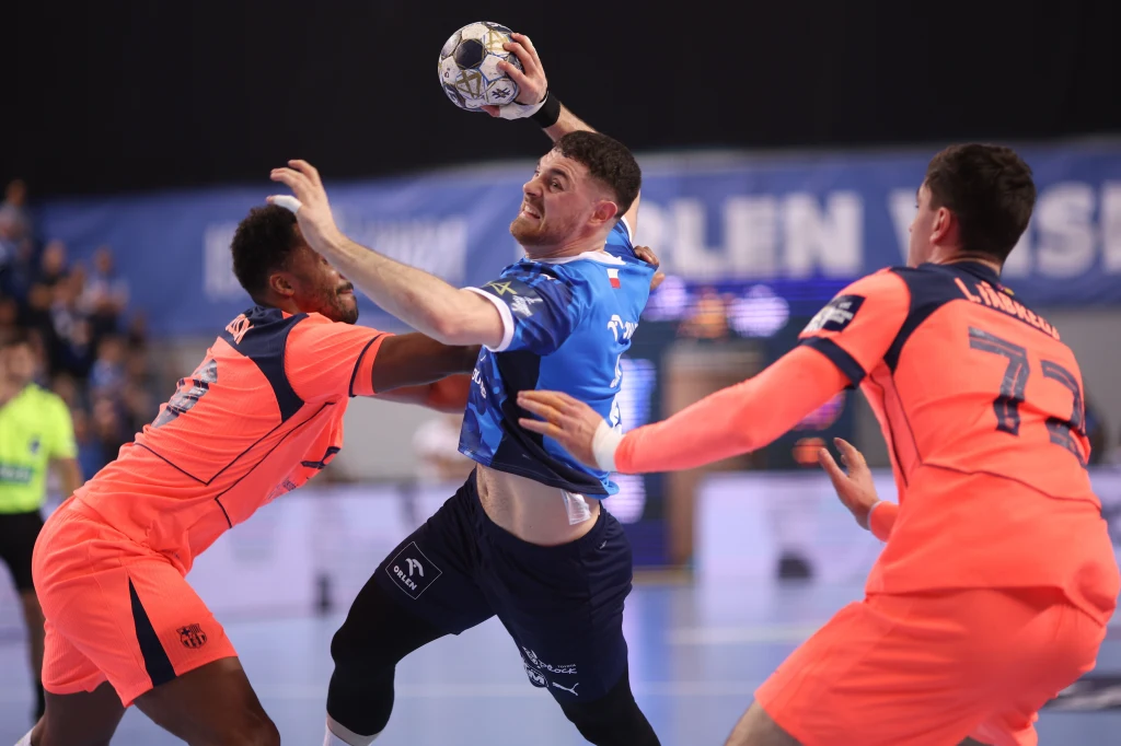 Trzech zawodników podczas dynamicznej akcji w meczu piłki ręcznej, jeden z nich w niebieskim stroju próbuje oddać rzut, podczas gdy dwóch rywali w pomarańczowych koszulkach stara się go powstrzymać.