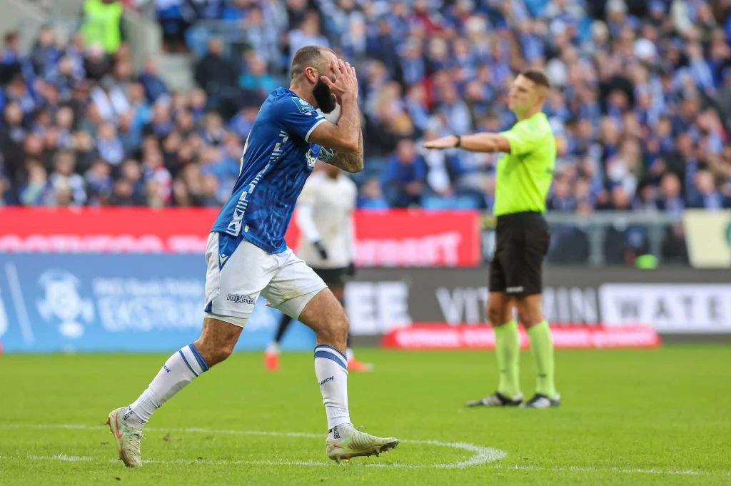 Piłkarz w niebieskiej koszulce zasłania twarz rękoma wyrażając emocje, w tle sędzia gestem sygnalizuje decyzję, a trybuny wypełnione są kibicami.