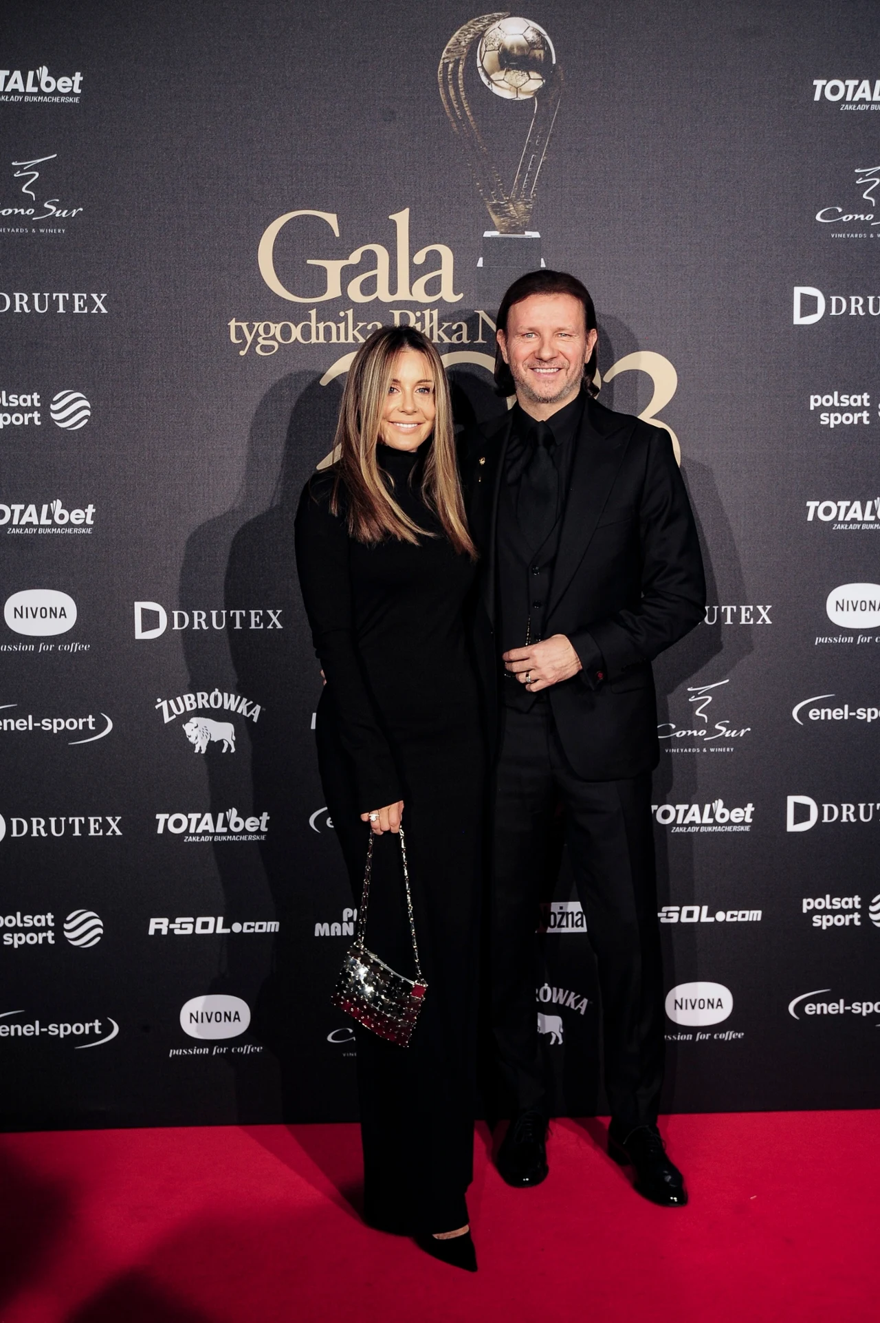 Radosław Majdan z małżonką Małgorzatą na salonach Para ubrana elegancko pozuje na czerwonym dywanie podczas gali piłkarskiej, na tle ścianki z logotypami sponsorów i grafiki wydarzenia.