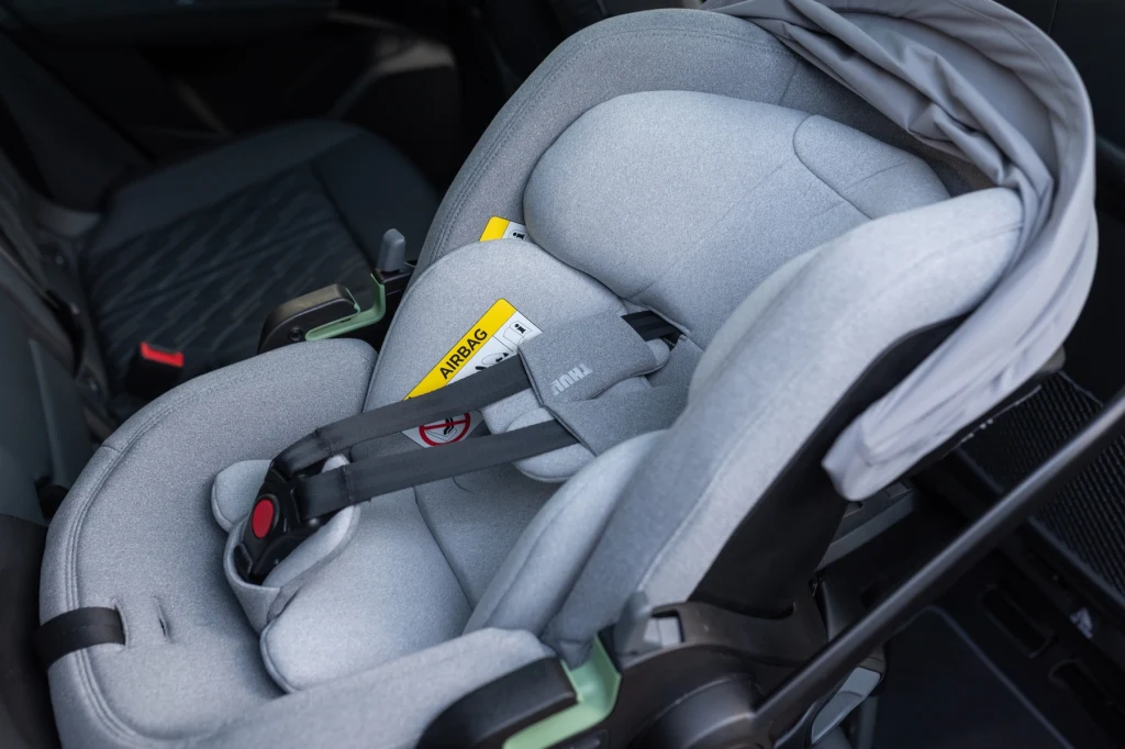Fotelik dziecięcy Thule Maple zamontowany w samochodzie, wyposażony w pasy bezpieczeństwa oraz specjalną naklejkę ostrzegającą przed poduszką powietrzną. Jasnoszara tapicerka, nowoczesny wygląd i funkcjonalność, wnętrze auta utrzymane w ciemnym kolorze.