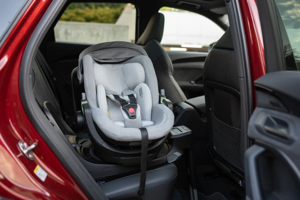 Fotelik dziecięcy Thule Maple zamontowany na tylnym siedzeniu samochodu osobowego, widoczny z otwartych bocznych drzwi pojazdu. Jasnoszare obicie fotelika, wyraźne pasy bezpieczeństwa oraz nowoczesne wnętrze auta.
