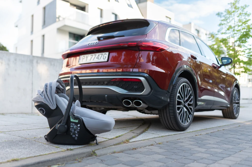 Czerwony samochód osobowy marki Audi zaparkowany na tle nowoczesnych budynków mieszkalnych. Na chodniku tuż za pojazdem ustawiony jest szary fotelik dziecięcy do samochodu.