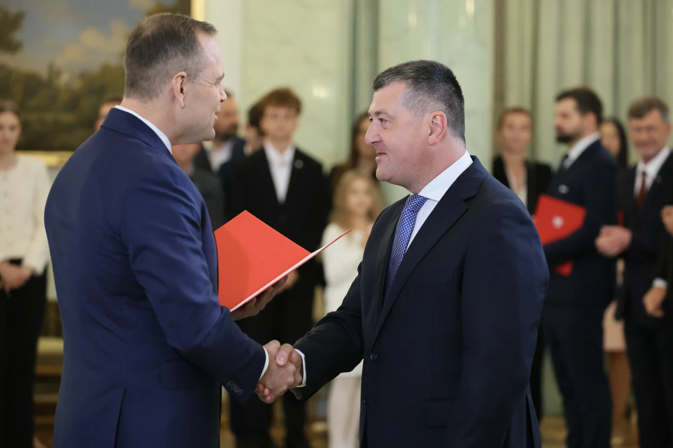 Leszek Skiba, doradca Karola Nawrockiego mówi wprost: jest przestrzeń w budżecie na realizację propozycji prezydenta dot. podniesienia progu podatkowego Dwóch mężczyzn w garniturach ściska sobie dłonie podczas oficjalnej ceremonii wręczenia dokumentów, w tle grupa osób ubranych elegancko obserwujących wydarzenie.