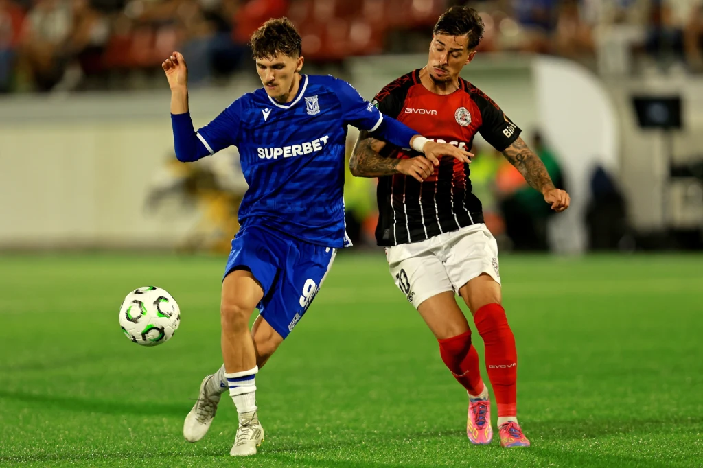 Mecz Lech Poznań - Lincoln Red Imps Dwóch piłkarzy rywalizuje o piłkę na boisku podczas meczu piłkarskiego, jeden ubrany w niebieski strój, drugi w czarno-czerwony; w tle widoczni kibice na trybunach.