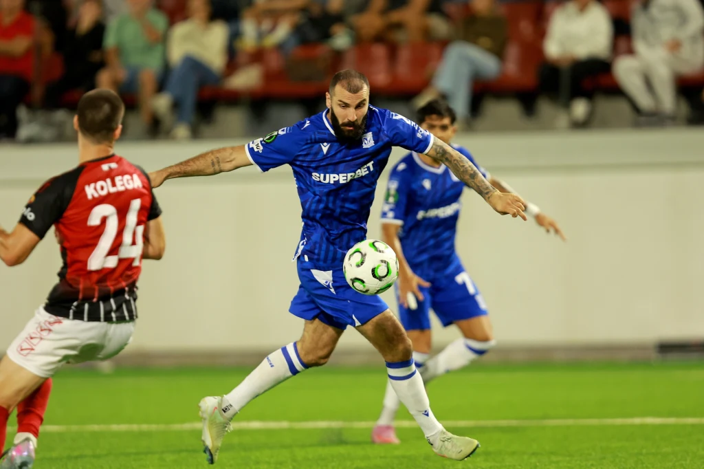 Mikael Ishak w meczu z Lincoln Red Imps Piłkarz w niebieskim stroju kopie piłkę na boisku podczas meczu, zbliżenie na dynamiczną akcję z udziałem zawodnika drużyny przeciwnej w czerwono-czarnym stroju, widownia w tle.