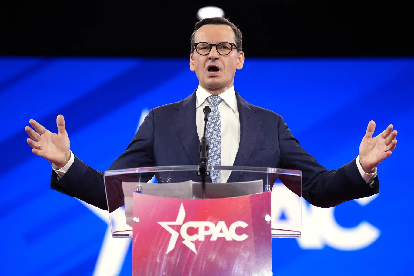 Mężczyzna w garniturze stoi za mównicą z logo CPAC, przemawia na tle niebieskiego ekranu, gestykulując rękami. W tle pojawia się charakterystyczny biały napis oraz logo wydarzenia.