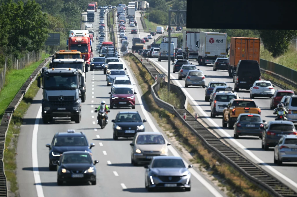 Korek samochodowy na autostradzie, liczne pojazdy w obu kierunkach, w tym ciężarówki, samochody osobowe i motocykliści między pasami, puste tory kolejowe po prawej stronie.