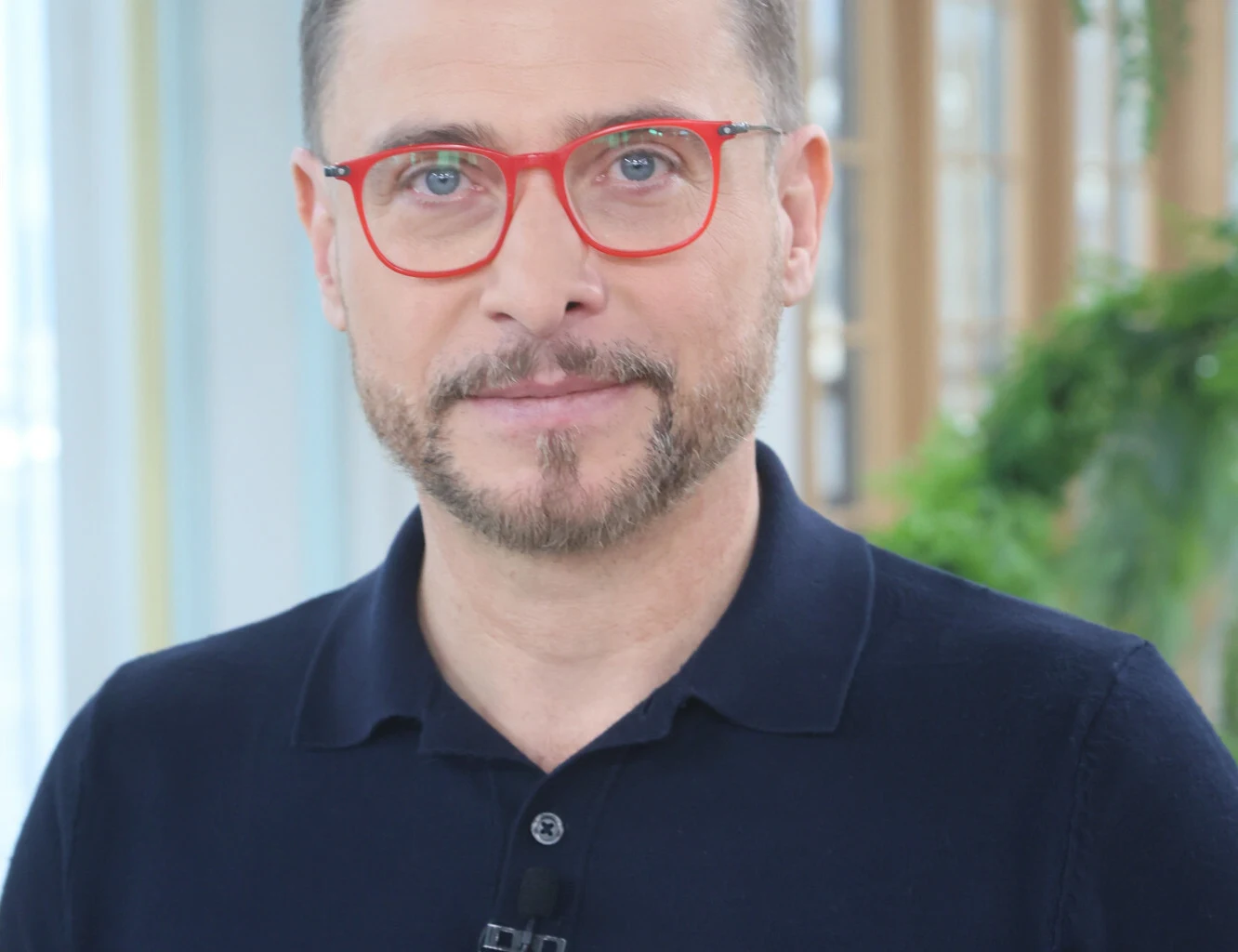 Mężczyzna w okularach o czerwonych oprawkach i krótkiej brodzie stoi w jasnym pomieszczeniu z zielenią w tle. Ubrany w ciemnogranatową koszulkę polo, patrzy bezpośrednio w obiektyw z delikatnym uśmiechem na twarzy.