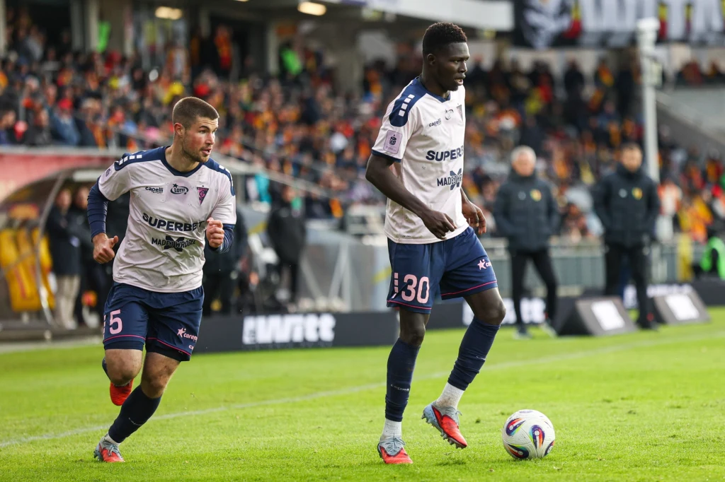 Kryspin Szcześniak i Ousmane Sow w barwach Górnika Zabrze Dwóch piłkarzy drużyny w białych koszulkach z numerami 25 i 30 rywalizuje na murawie stadionu, piłka znajduje się przy stopie zawodnika oznaczonego numerem 30, w tle trybuny wypełnione kibicami.