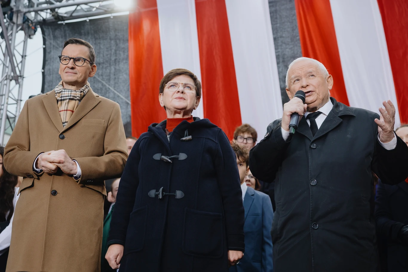 Jarosław Kaczyński i byli premierzy: Mateusz Morawiecki i Beata Szydło podczas manifestacji przeciwko imigracji Trzy osoby w eleganckich płaszczach stoją przed dużą biało-czerwoną flagą, jedna z nich mówi do mikrofonu, pozostali przysłuchują się uważnie. W tle zgromadzenie ludzi, atmosfera oficjalna.