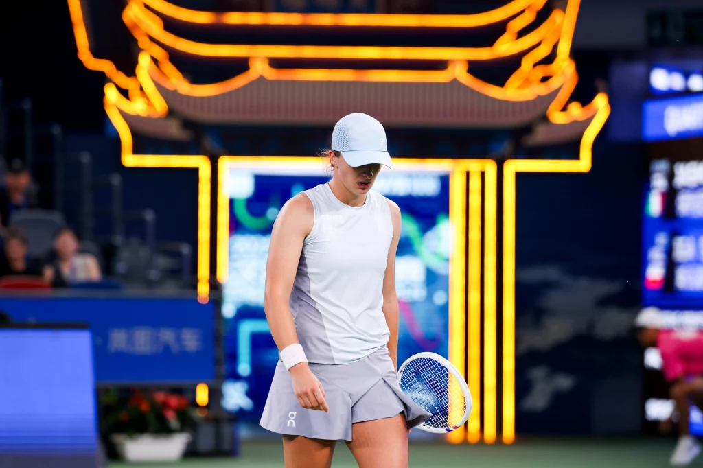 Zawodniczka tenisowa w sportowym stroju stoi na korcie z rakietą w ręku, w tle widoczne są jasne, neonowe dekoracje przypominające tradycyjną architekturę azjatycką oraz elektroniczna tablica.