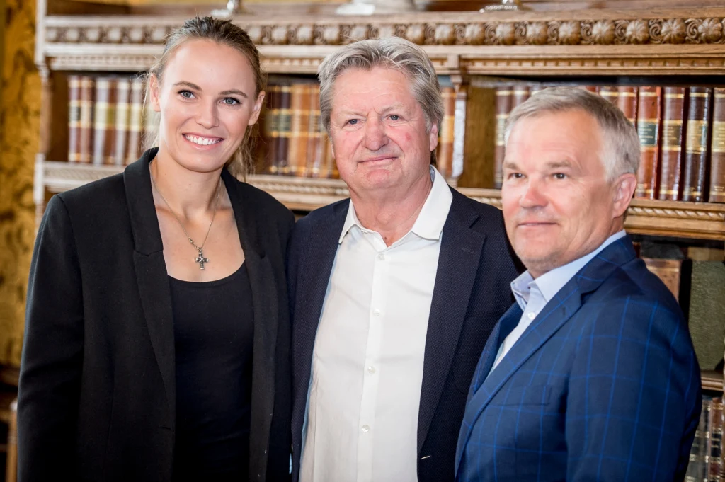 Troje elegancko ubranych osób pozuje razem na tle regału z książkami, wszyscy uśmiechnięci i patrzący w stronę aparatu.