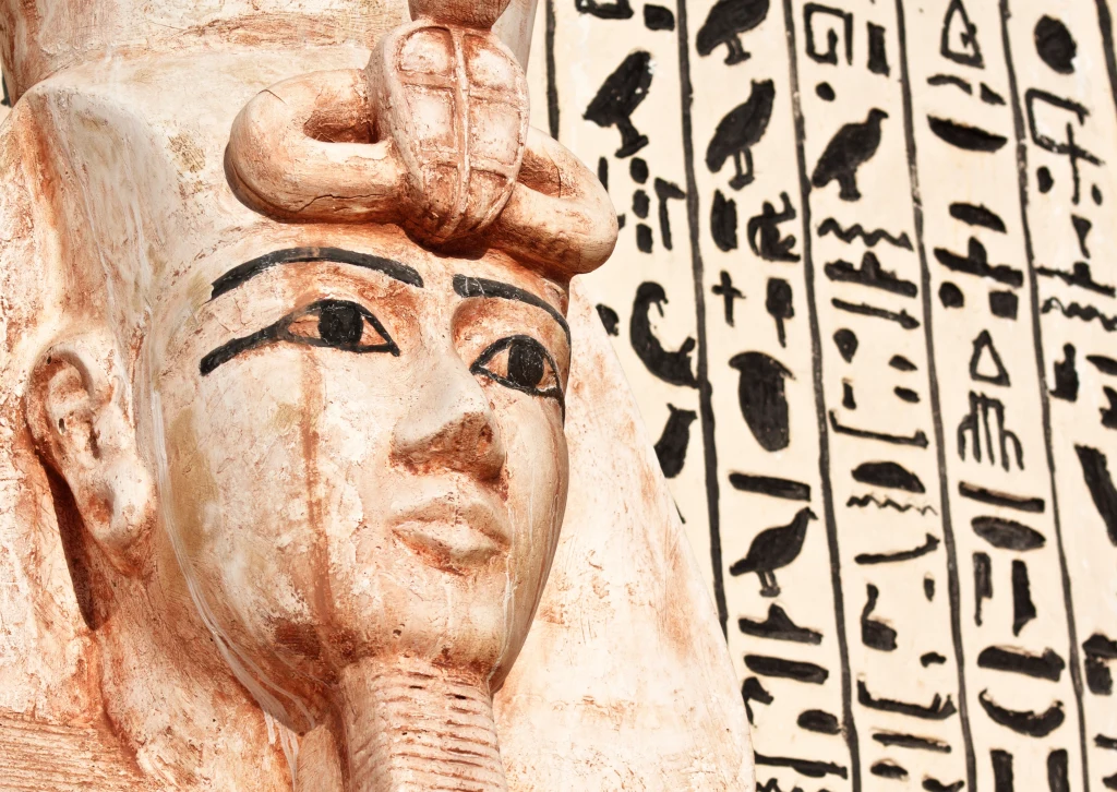 Główna część kadru wypełniona jest szczegółową rzeźbą egipskiego faraona wyrzeźbioną w kamieniu, po lewej stronie widoczne są wyraźne detale twarzy i tradycyjny nemes na głowie. W tle znajduje się kamienna ściana pokryta pionowymi kolumnami starożytnyc...