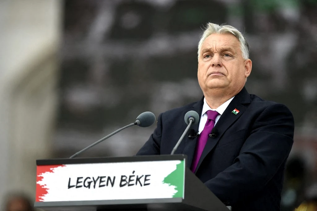 Viktor Orban planuje obejść sankcje USA na rosyjskie koncerny naftowe Mężczyzna w garniturze i fioletowym krawacie stoi za mównicą z napisem 'LEGYEN BÉKE' oraz flagą w barwach Węgier, w tle rozmyte tło, dominujące kolory to szarość i biel.