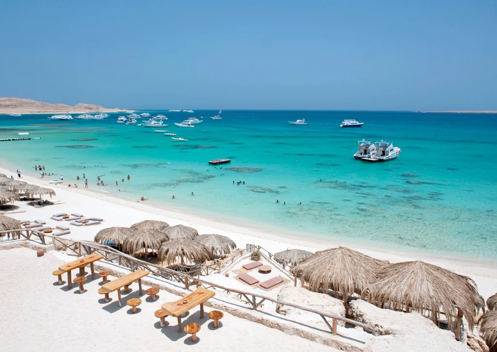 Plaża z białym piaskiem, turkusowa woda, liczne łodzie i jachty cumujące przy brzegu, ludzie kąpiący się w wodzie i korzystający z wypoczynku, drewniane stoły oraz parasole z liści palmowych tworzące strefę relaksu.