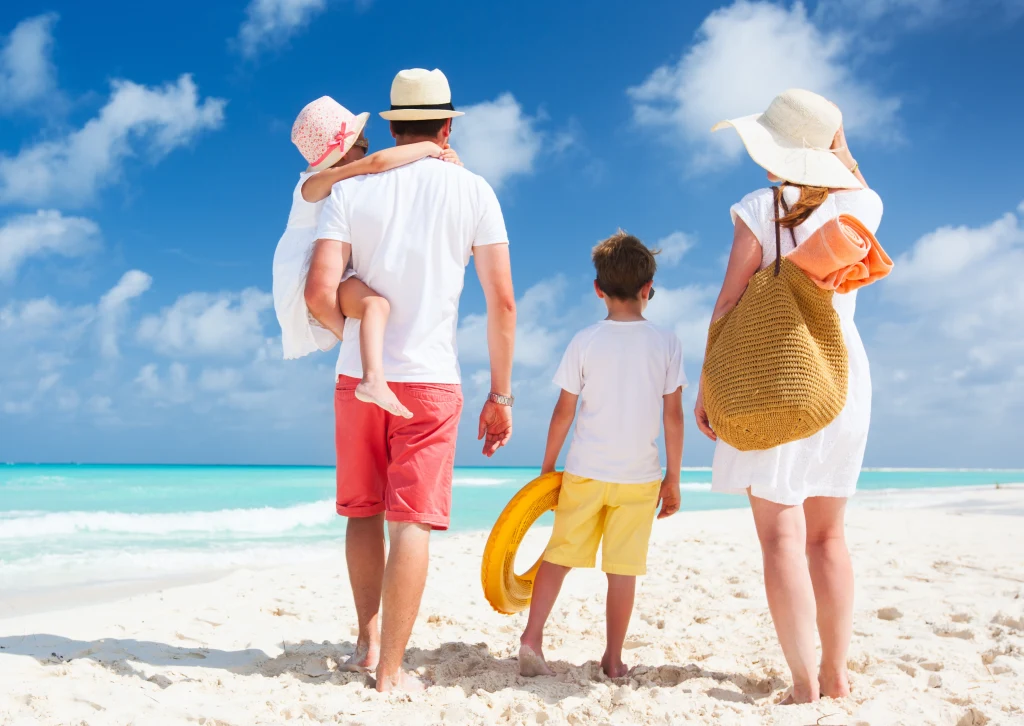 Rodzina spaceruje brzegiem morza po piaszczystej plaży, rodzice i dwójka dzieci ubrani są w letnie ubrania, wszyscy odwróceni plecami do aparatu, a słońce świeci na błękitnym niebie z delikatnymi chmurami.