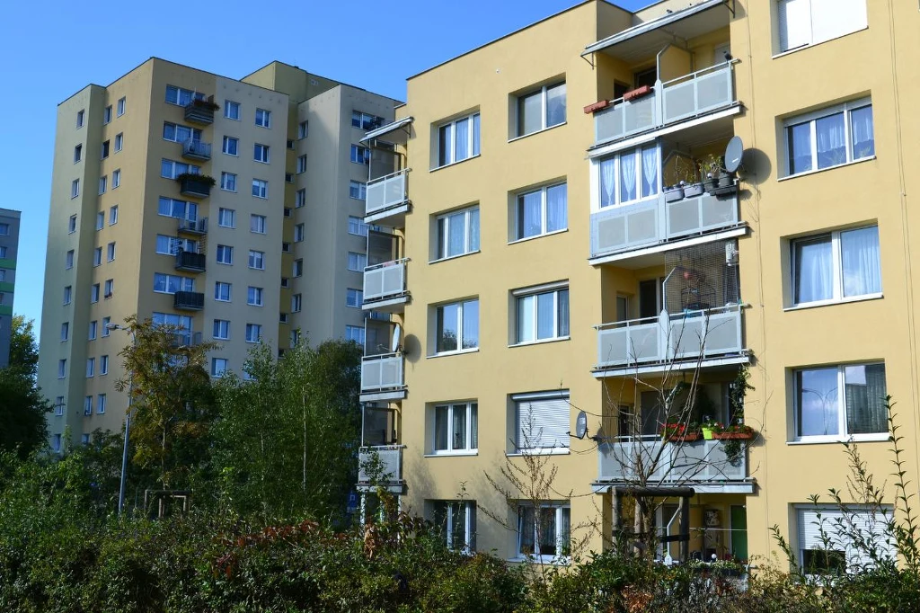 Brak windy w bloku lub kamienicy zdecydowanie wpływa na pogorszenie się jakości życia osób zmagających się z różnymi niepełnosprawnościami Bloki mieszkalne z żółtymi elewacjami, szerokimi balkonami i licznymi antenami satelitarnymi, otoczone zielenią i drzewami w słoneczny dzień.