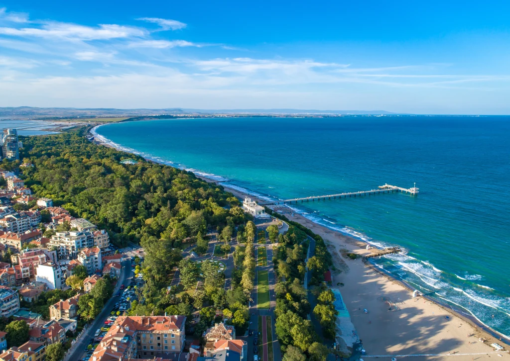 Panorama nadmorskiego miasta z szeroką plażą, długim molo wchodzącym w morze, zielonym parkiem pełnym drzew oraz zabudową miejską rozciągającą się wzdłuż wybrzeża.