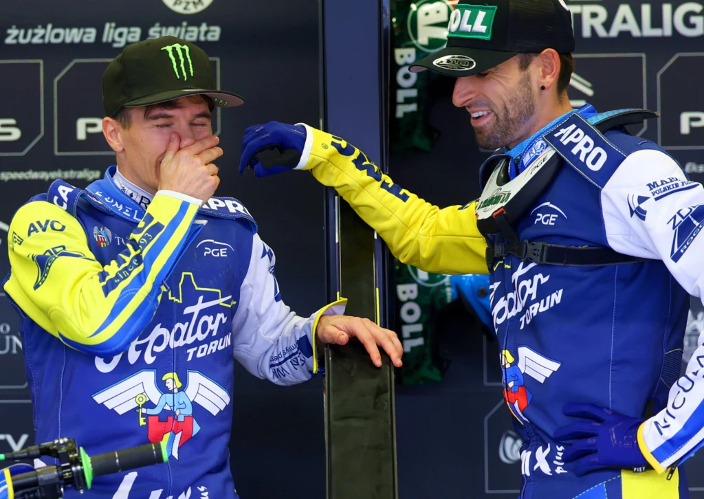 Dwóch żużlowców w niebiesko-żółtych kombinezonach drużyny Apator Toruń stoi w boksach, jeden z nich śmieje się i zakrywa twarz dłonią, drugi klepie go przyjaźnie po ramieniu.