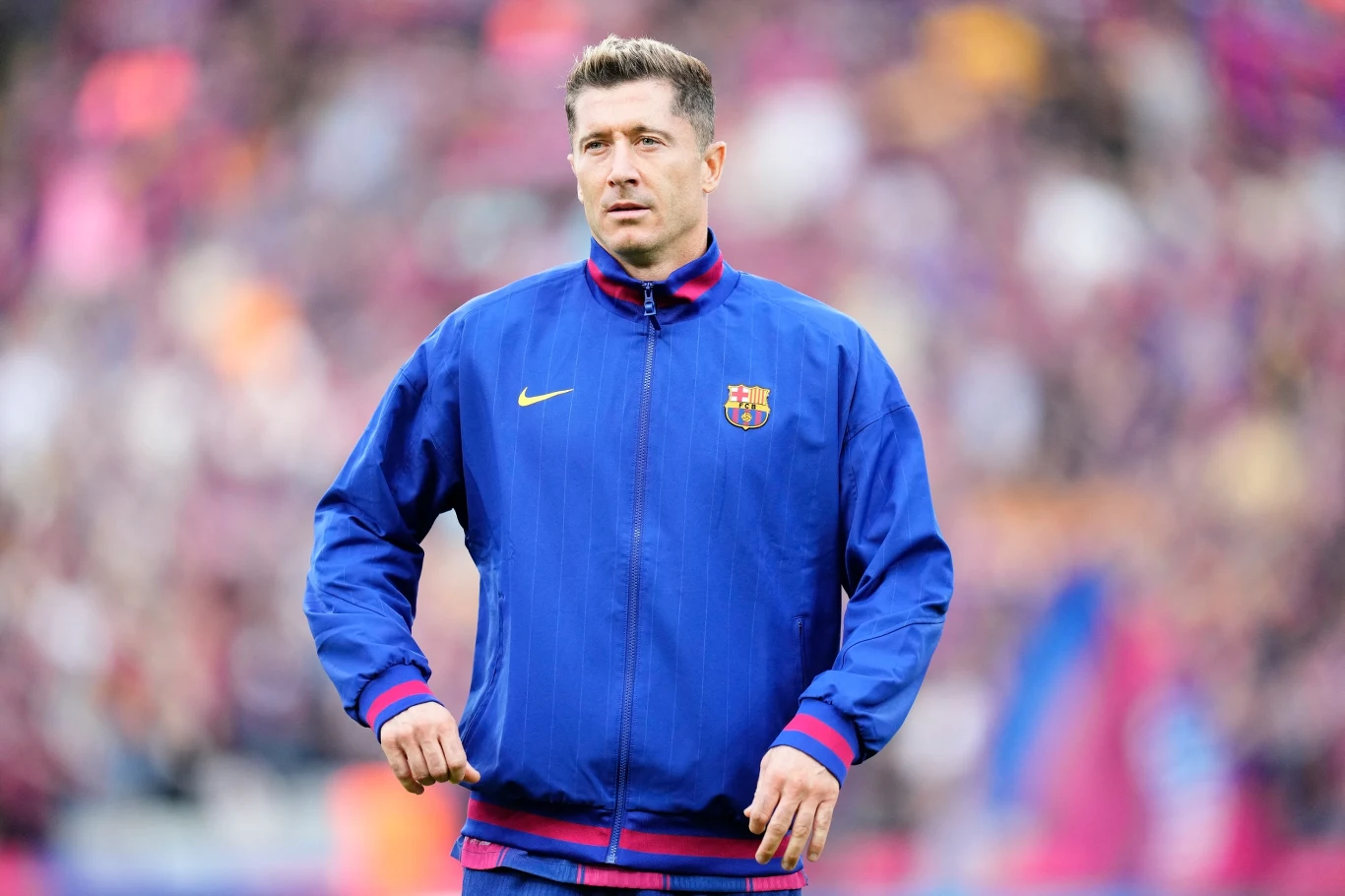 Robert Lewandowski Piłkarz w niebieskiej kurtce sportowej z herbem FC Barcelony na tle stadionu, widoczne rozmyte barwy kibiców na trybunach.